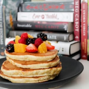 Stack of pancakes topped with fresh berries and oranges against a backdrop of books.