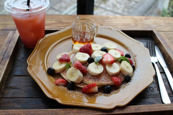 Mouth-watering banana and berry pancakes served with syrup and juice on a wooden table.